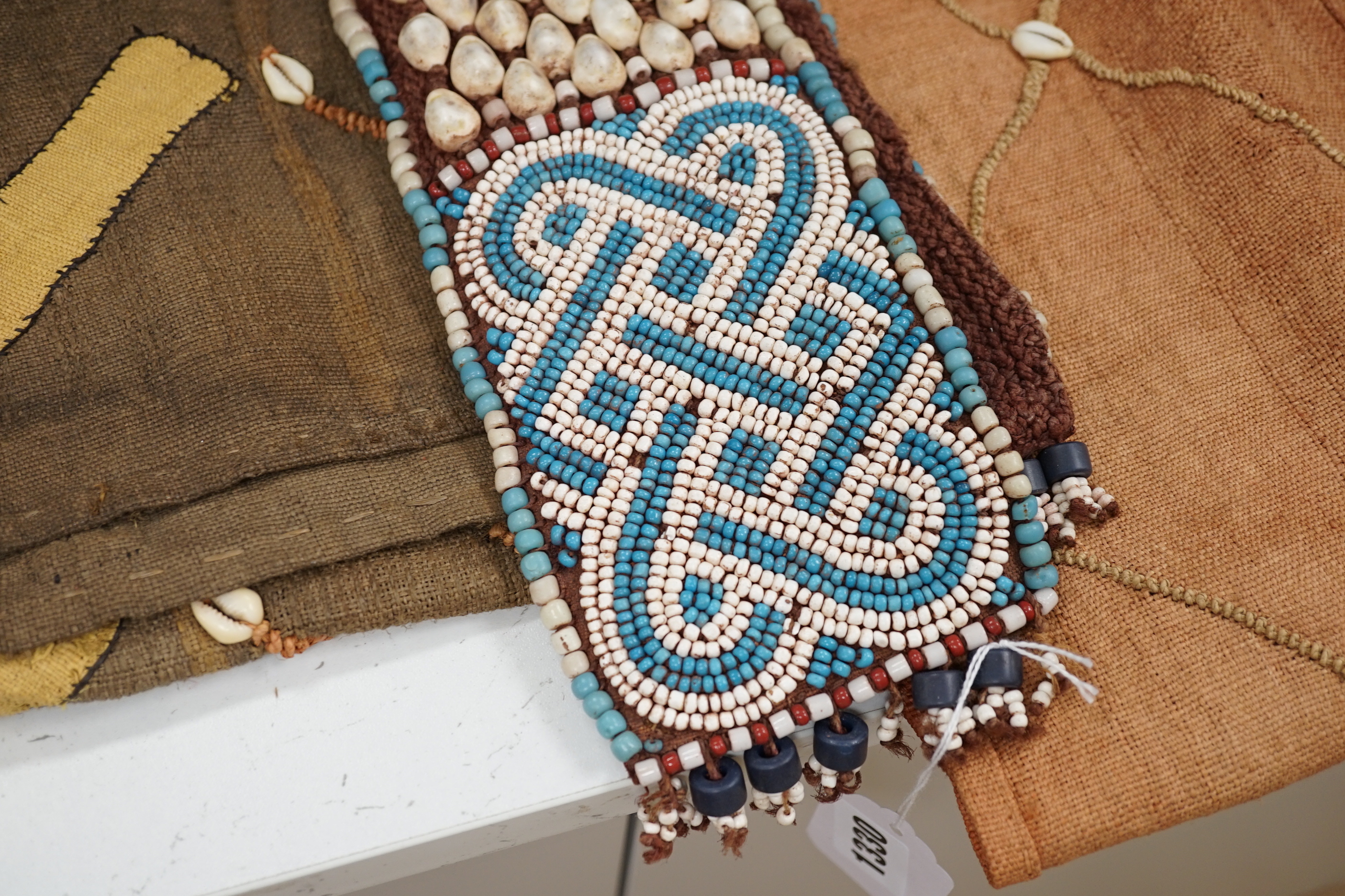 Three African tribal hangings, embroidered with coloured hand dyed threads and appliquéd shells on smaller handwoven cloth panels all sewn together to form the hangings, including a chieftains coloured bead worked and sh